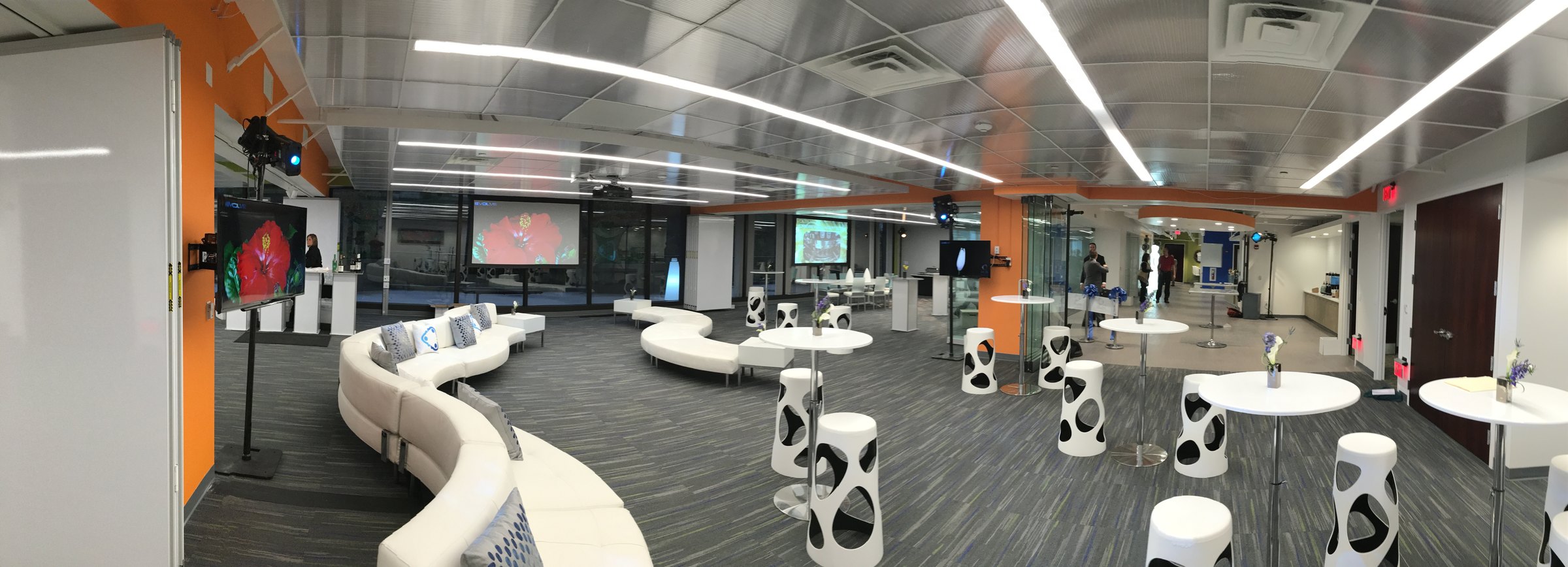 Panoramic view of a corporate hospitality lounge with multiple wall-mounted displays, projection screens, accent uplighting, modular lounge seating and a catering station