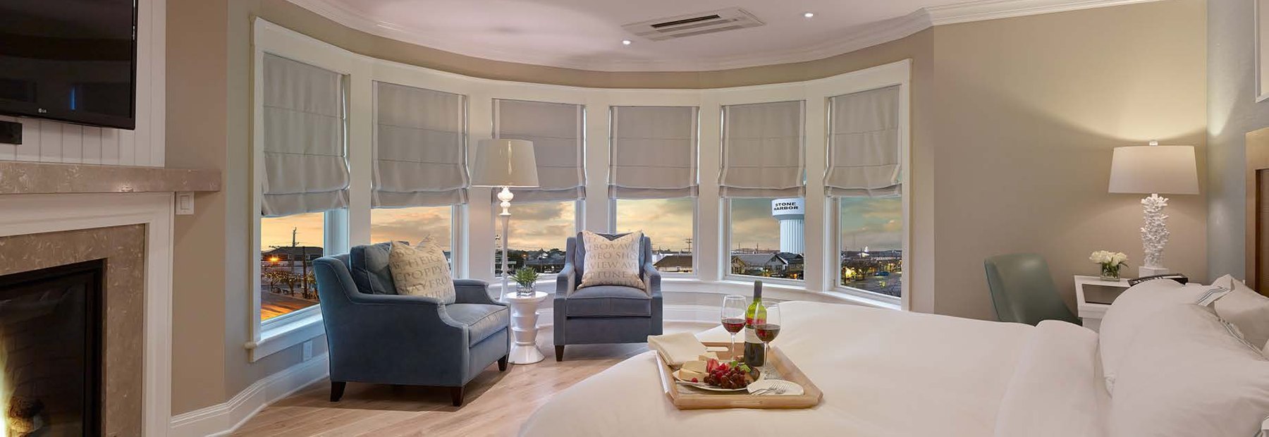 Guest suite with bay window seating, wall-mounted display above a fireplace, and layered window treatments
